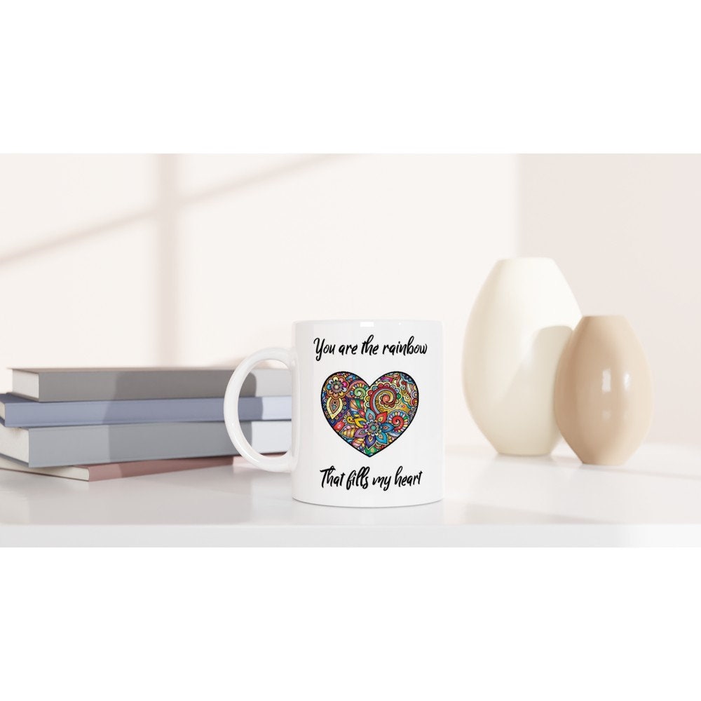 A white ceramic mug on a table with the words "You are the rainbow that fills my heart" in black and a picture of a hear with multicoloured rainbow intricate designs.