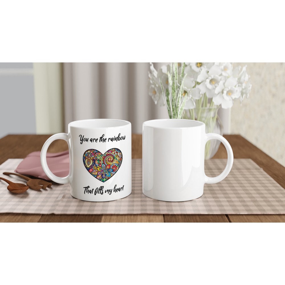The front and back view of a white ceramic mug with the words "You are the rainbow that fills my heart" in black and a picture of a hear with multicoloured rainbow intricate designs.