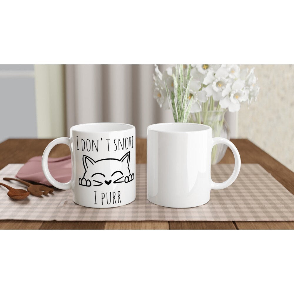 The front and back views of a white ceramic mug with the words "I don't snore, I purr" and an image of a cat peeping up happily, in black