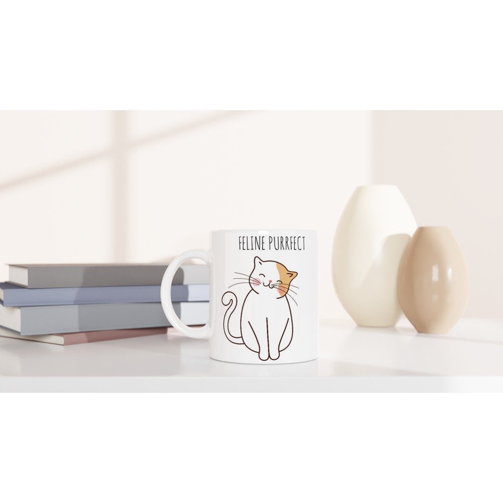 A white ceramic mug on a table with the words "Feline purrfect" in black and a picture of a white and light brown cat smiling and sitting happily.