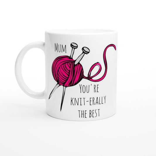 A white ceramic mug with the words, "Mum, you're knit-erally the best" in black and a picture of a pink ball of yarn with two knitting needles stuck through it.