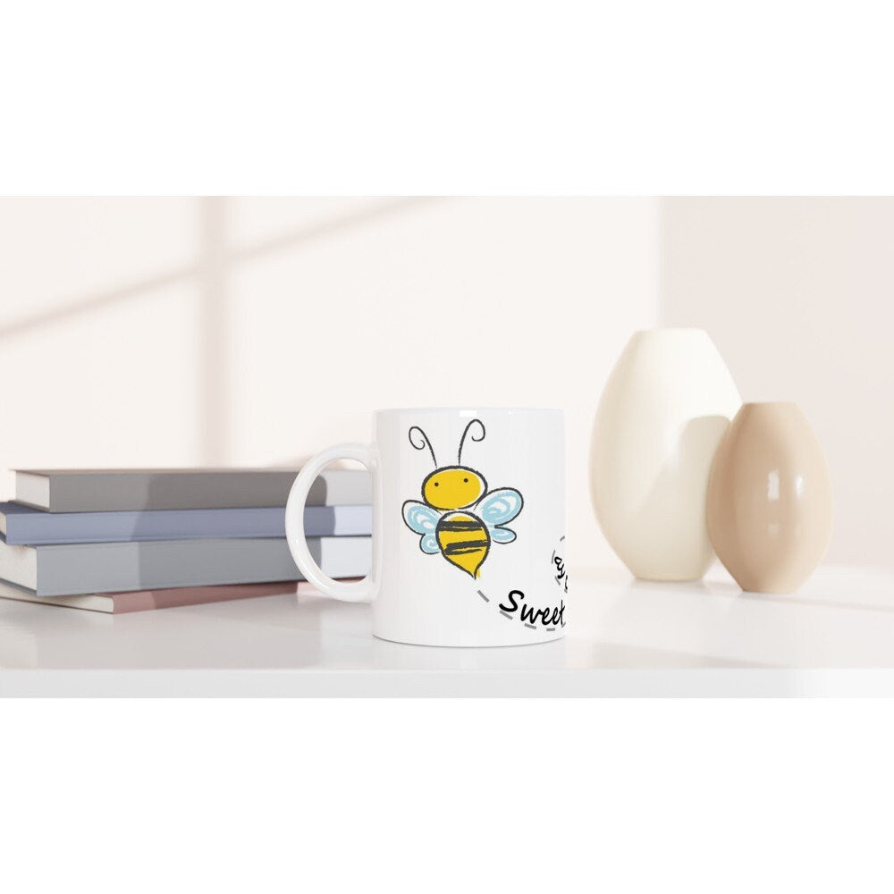 A white ceramic mug on a table with black text saying, "Sweet as can bee" and the image of a drawing of a bee and it's path across the cup.