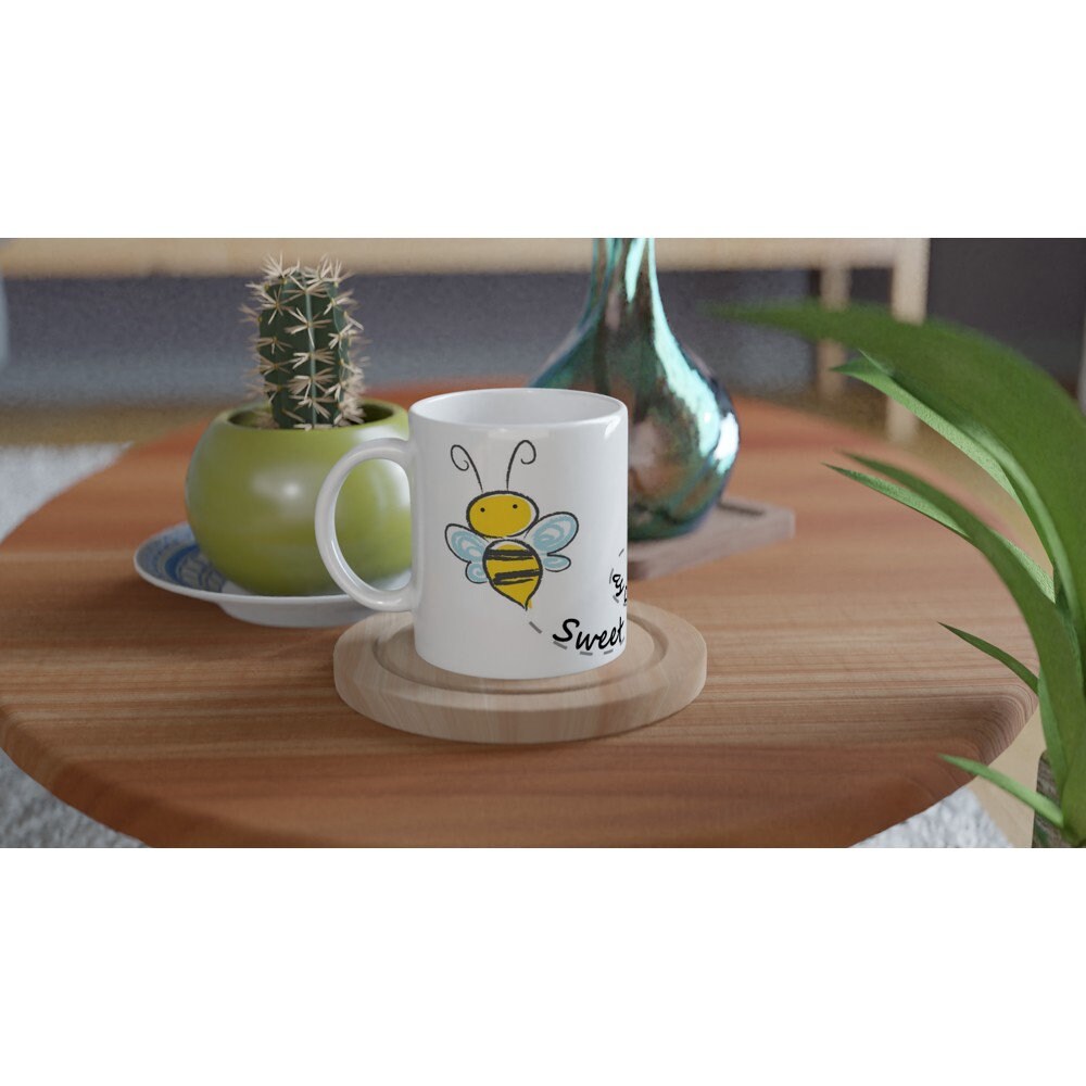 A white ceramic mug on a wooden table with black text saying, "Sweet as can bee" and the image of a drawing of a bee and it's path across the cup.