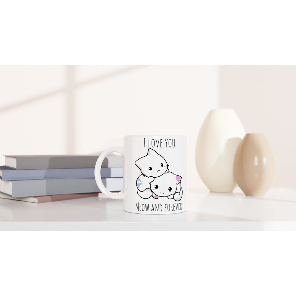 A white ceramic mug on a table with the pun, "I love you meow and forever" and a picture of a blue and white striped cat next to a white and pink spotted cat.