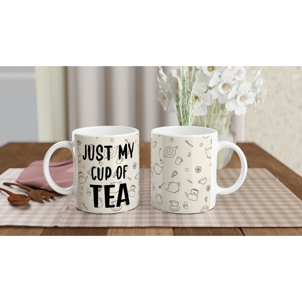 The front and back views of a white ceramic mug with bold black text saying, "Just my cup of tea" and line drawings of tea themed items brown on an off white background.