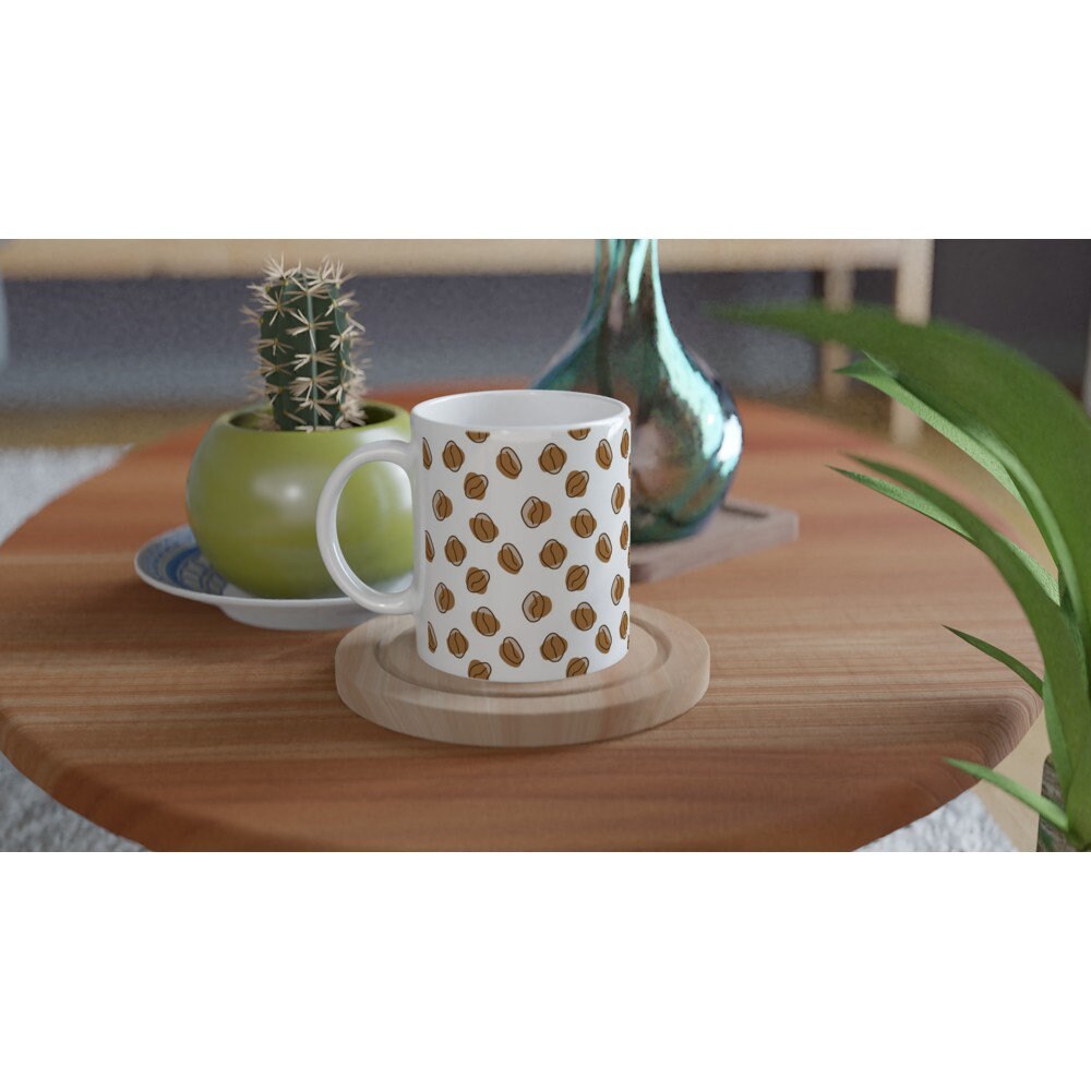 White ceramic mug with a pattern of abstract brown coffee beans all over it, on a table with a cactus and vase.