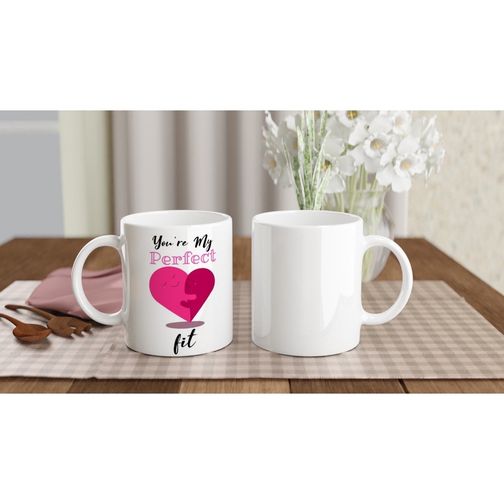 The front and back view of a white ceramic mug with the words, "you're my perfect fit" written on it and am image of a pink hear made of two halves which are facing each other, smiling and hugging.