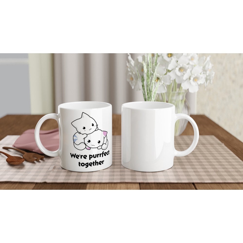 A white ceramic mug on a table with the words, "we're purrfect together" and the image of a white and blue striped cat next to a white and pink spotted kitten