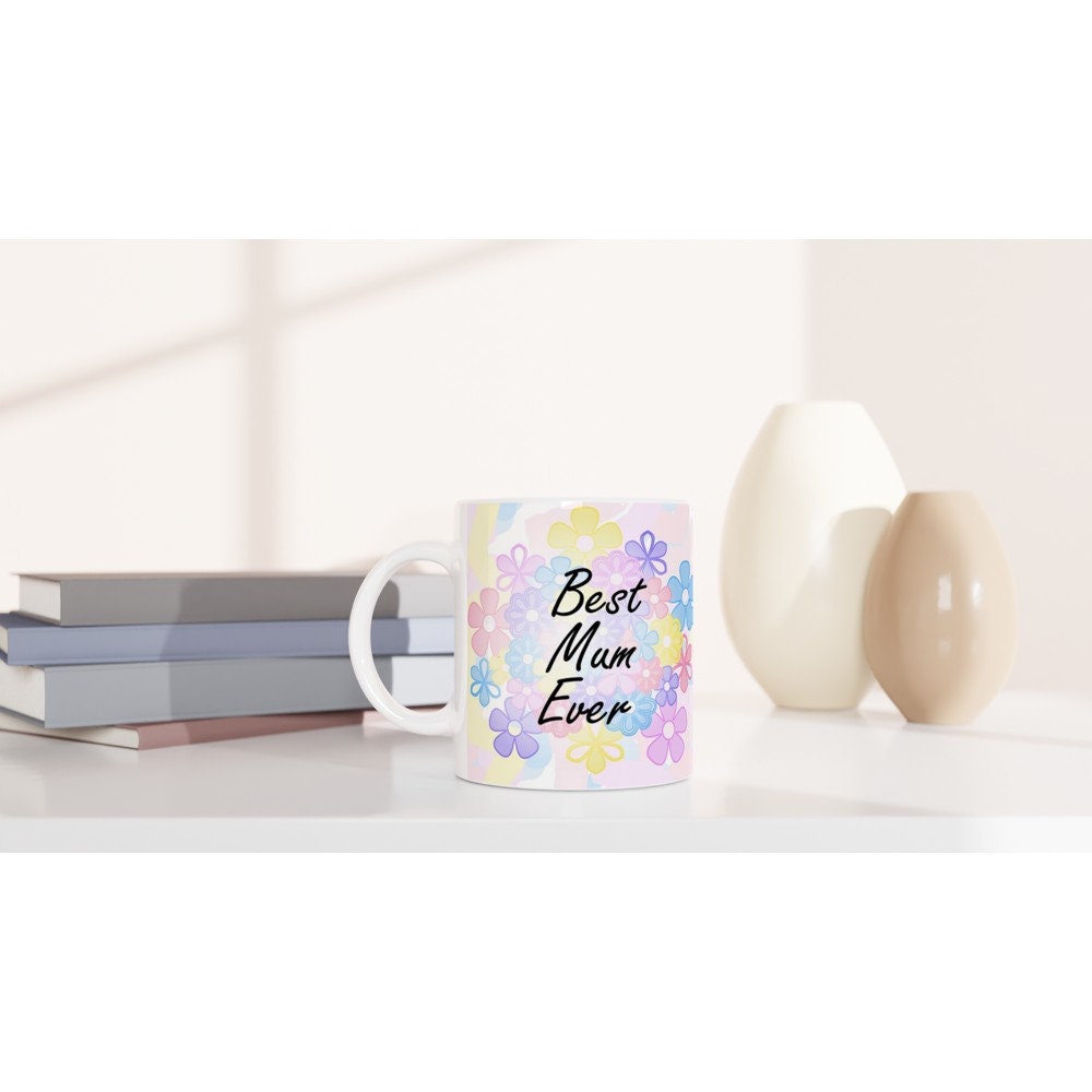 Front view of a white ceramic mug with the words "Best Mum Ever" written on it, with a background of pastel coloured flowers on a table.