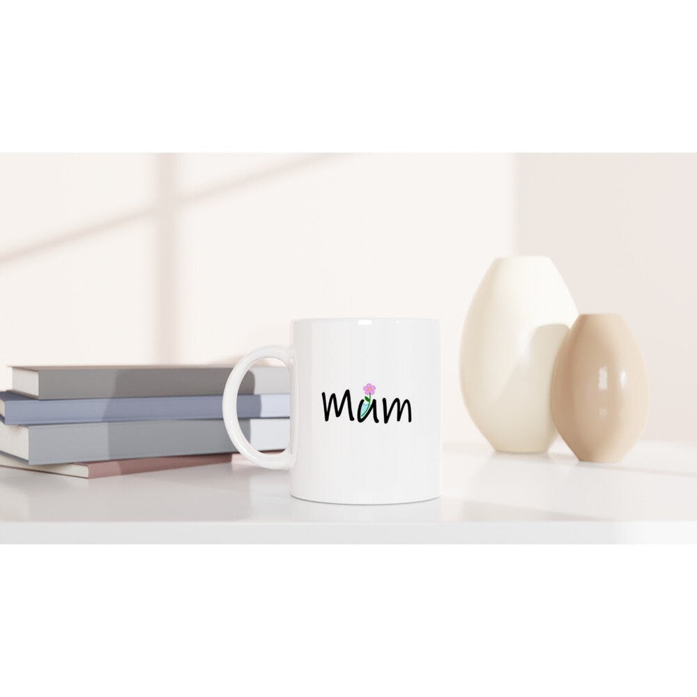 A white ceramic mug on a table with the word, "Mum" written on it in black where the letter "U" is a container with water in it and a pink flower sticking out.