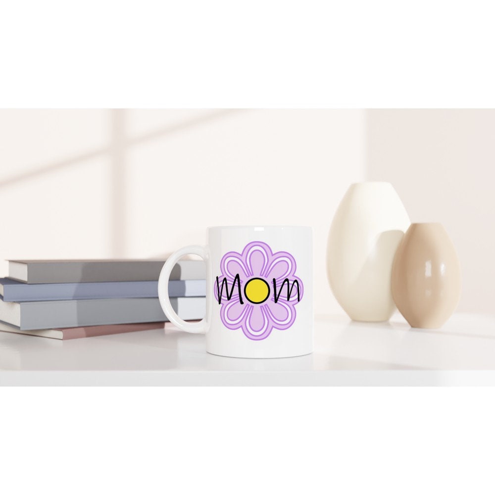 A white ceramic mug on a table with the words "Mom" printed on it in black and a large purple flower in the background.