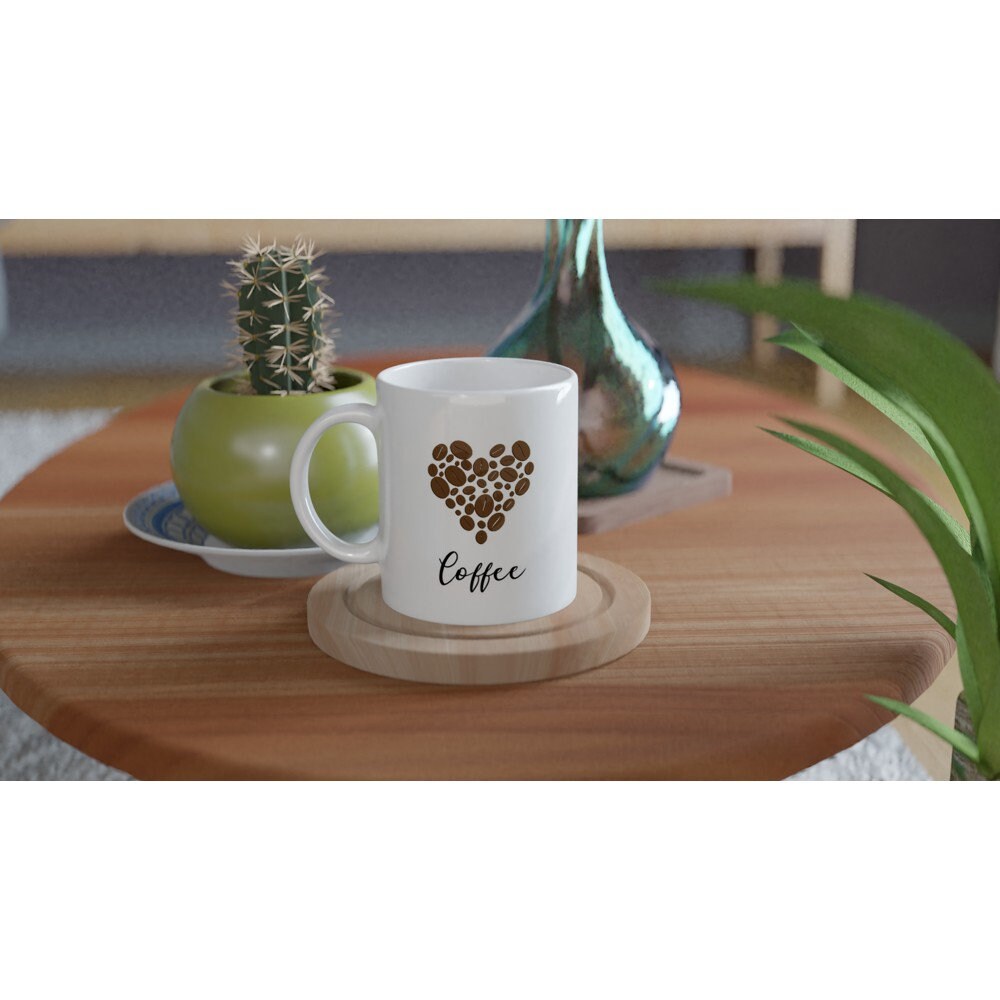 A white ceramic mug on a table with the word "coffee" in black and a picture of a heart made out of coffee beans above it.