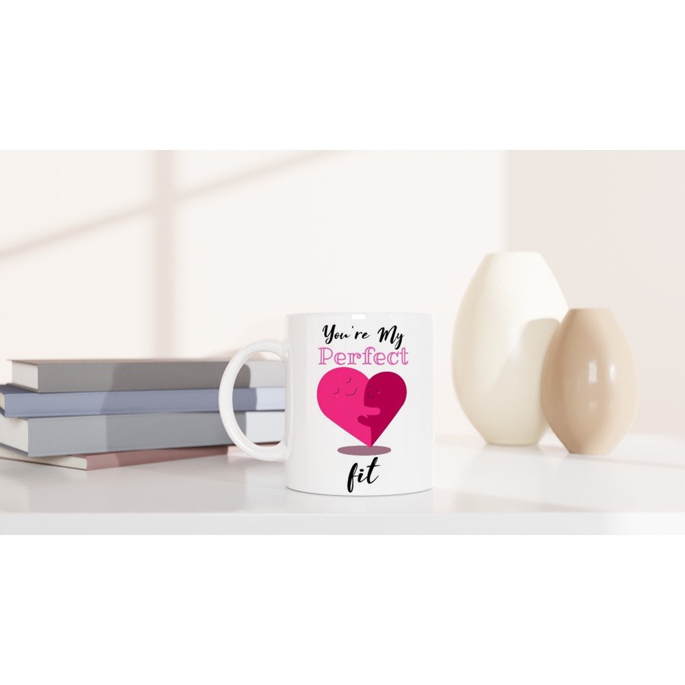 A white ceramic mug on a table with the words, "you're my perfect fit" written on it and am image of a pink hear made of two halves which are facing each other, smiling and hugging.